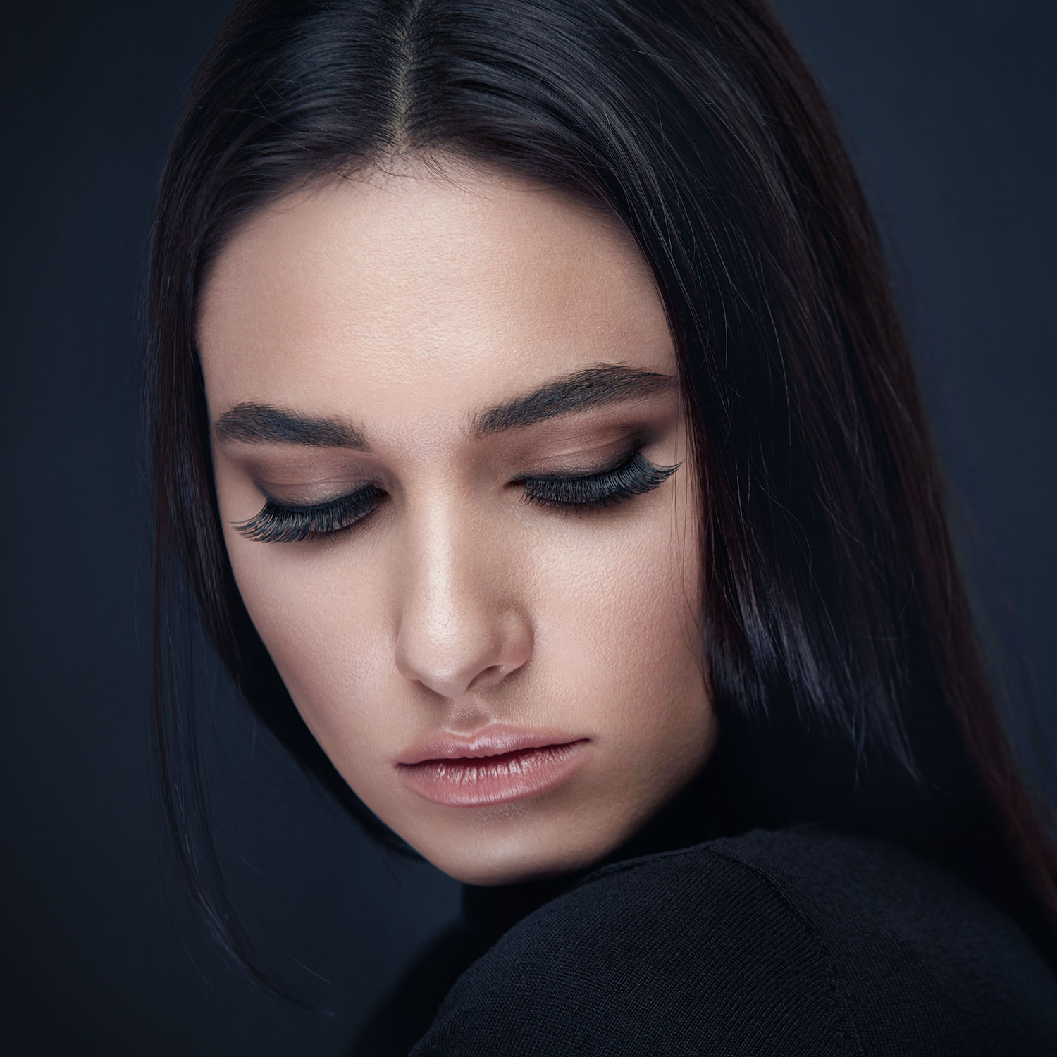 Thick, black hair and sculpted eyebrows frame beautiful, thick eyelashes. The woman is pictured on a dark background wearing a black shirt and has natural makeup and bronzed skin.